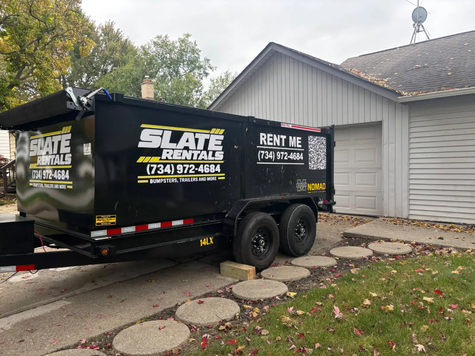 Slate Rentals dump trailer parked in a residential driveway next to a garage, with a wooden board under the tires to protect the concrete.