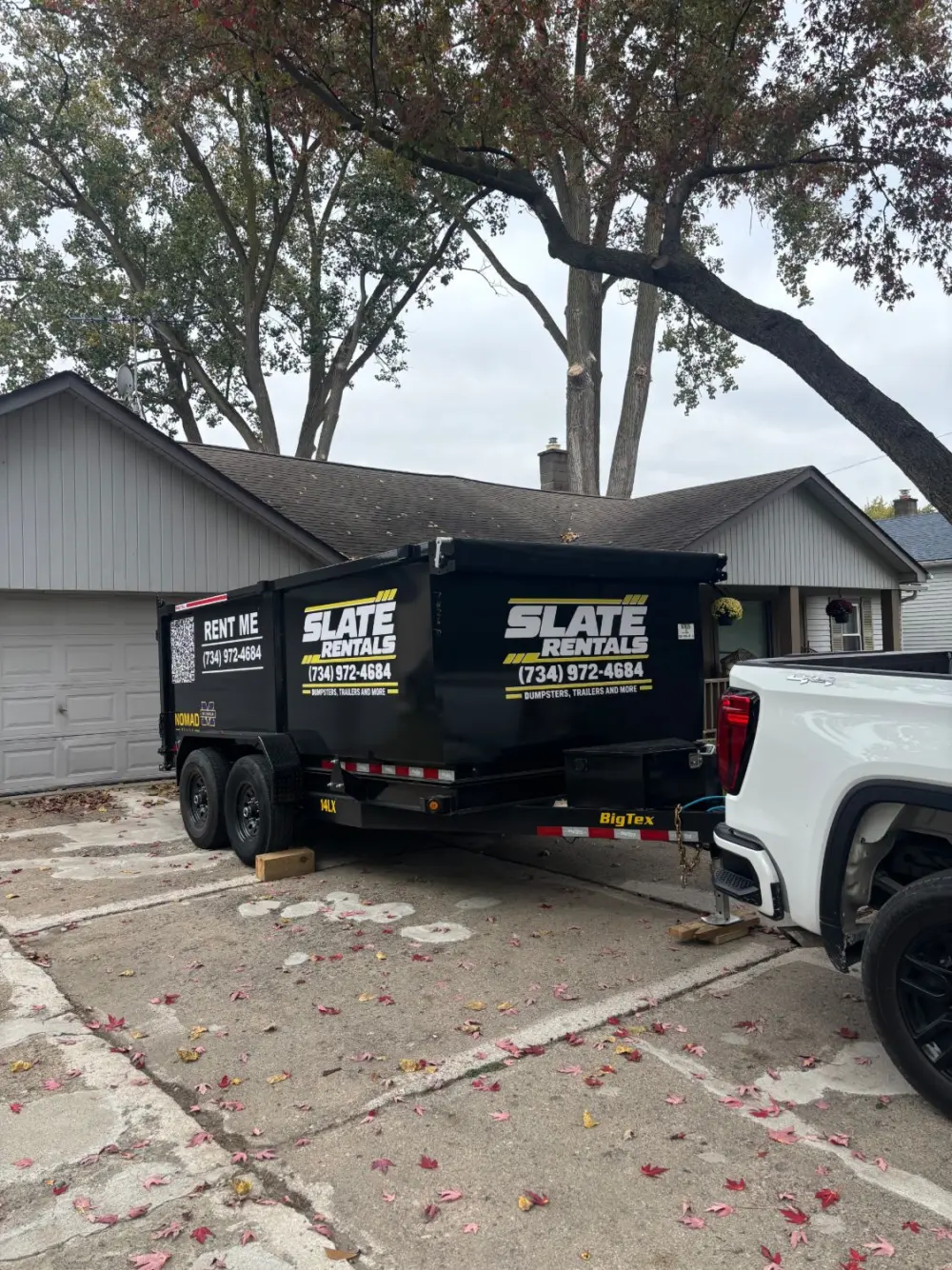 Dump trailer hitched to a white pickup in a driveway under a large maple tree.